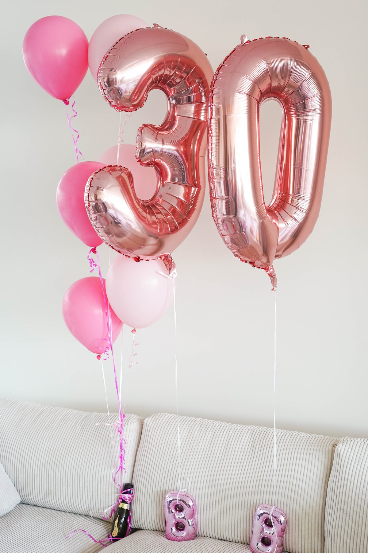 Pink and rose gold foil balloons, including large number 30 balloons and smaller pink heart balloons, floating above a beige sofa with small pink '8' balloons on the seat.